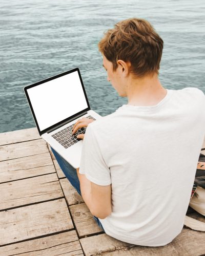 Jeune homme en tee-shirt blanc au bords de la mer, laptop en main en train de lire du texte sur les TVA en France